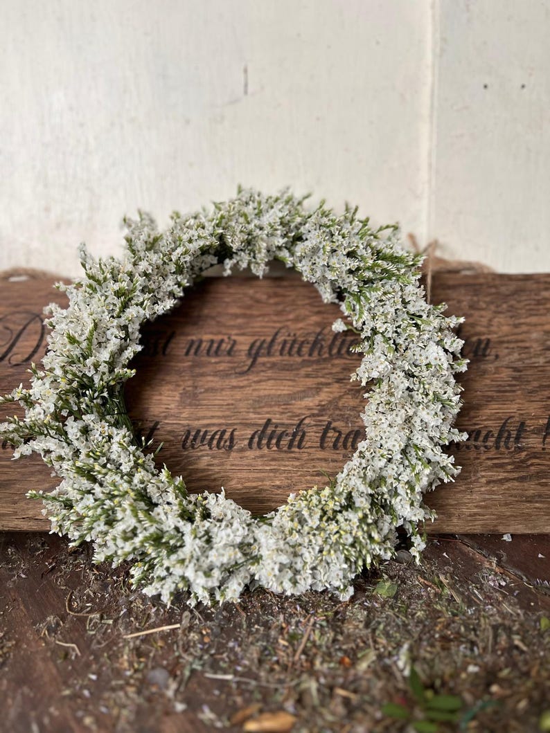 May include: A circular wreath composed of small white flowers and green stems. The wreath is presented on a wooden surface with a wooden sign in the background. The sign has text on it.