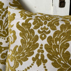 May include: Two cushions with a white backdrop and a repeating pattern in a golden-yellow hue. The pattern features a classic damask design with floral and leaf motifs.