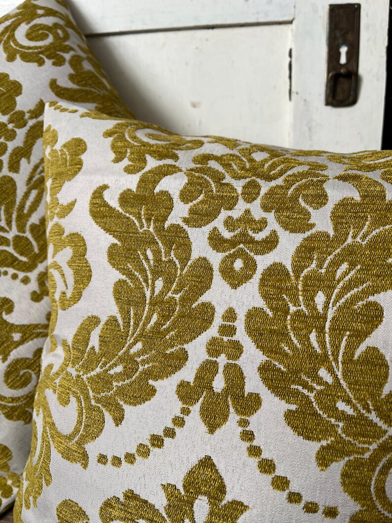 May include: Two cushions with a white backdrop and a repeating pattern in a golden-yellow hue. The pattern features a classic damask design with floral and leaf motifs.