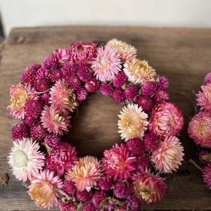 May include: A circular floral wreath composed of dried flowers in shades of pink, magenta, and peach. The wreath is densely packed with small, textured blooms, creating a full and vibrant appearance. The flowers are arranged in a circular shape, perfect for home decor.