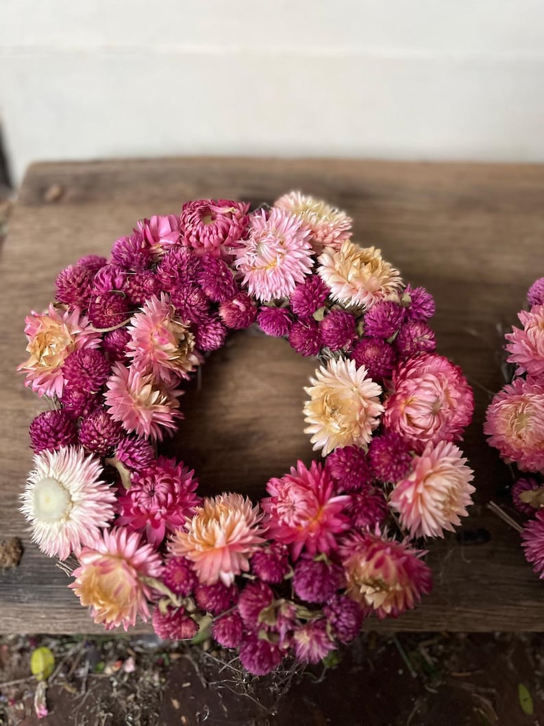 May include: A circular floral wreath composed of dried flowers in shades of pink, magenta, and peach. The wreath is densely packed with small, textured blooms, creating a full and vibrant appearance. The flowers are arranged in a circular shape, perfect for home decor.