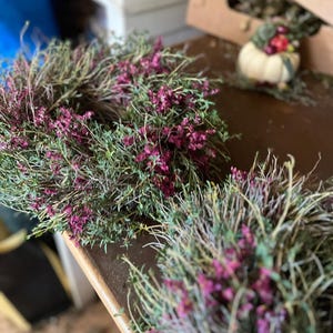 K&ouml;nnte beinhalten: Ein kreisf&ouml;rmiger Kranz aus getrockneten gr&uuml;nen Stielen und kleinen, leuchtend magentafarbenen Bl&uuml;ten. Der Kranz zeichnet sich durch eine dichte, strukturierte Anordnung nat&uuml;rlicher botanischer Elemente aus, die eine organische Komposition bilden.