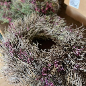 K&ouml;nnte beinhalten: Ein kreisf&ouml;rmiger Kranz aus getrockneten braunen Zweigen und &Auml;sten, der mit kleinen B&uuml;scheln leuchtend violetter Bl&uuml;ten durchsetzt ist, die in das strukturierte, holzige Material eingearbeitet sind. Der Kranz hat eine hohle Mitte.