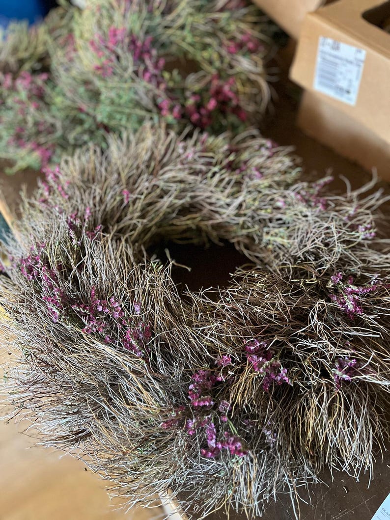 K&ouml;nnte beinhalten: Ein kreisf&ouml;rmiger Kranz aus getrockneten braunen Zweigen und &Auml;sten, der mit kleinen B&uuml;scheln leuchtend violetter Bl&uuml;ten durchsetzt ist, die in das strukturierte, holzige Material eingearbeitet sind. Der Kranz hat eine hohle Mitte.