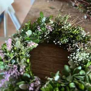 May include: A circular wreath constructed from green boxwood leaves, interspersed with small clusters of white baby's breath, light pink statice flowers, and spiky blue-green thistle heads, arranged in a dense, textured botanical ring.