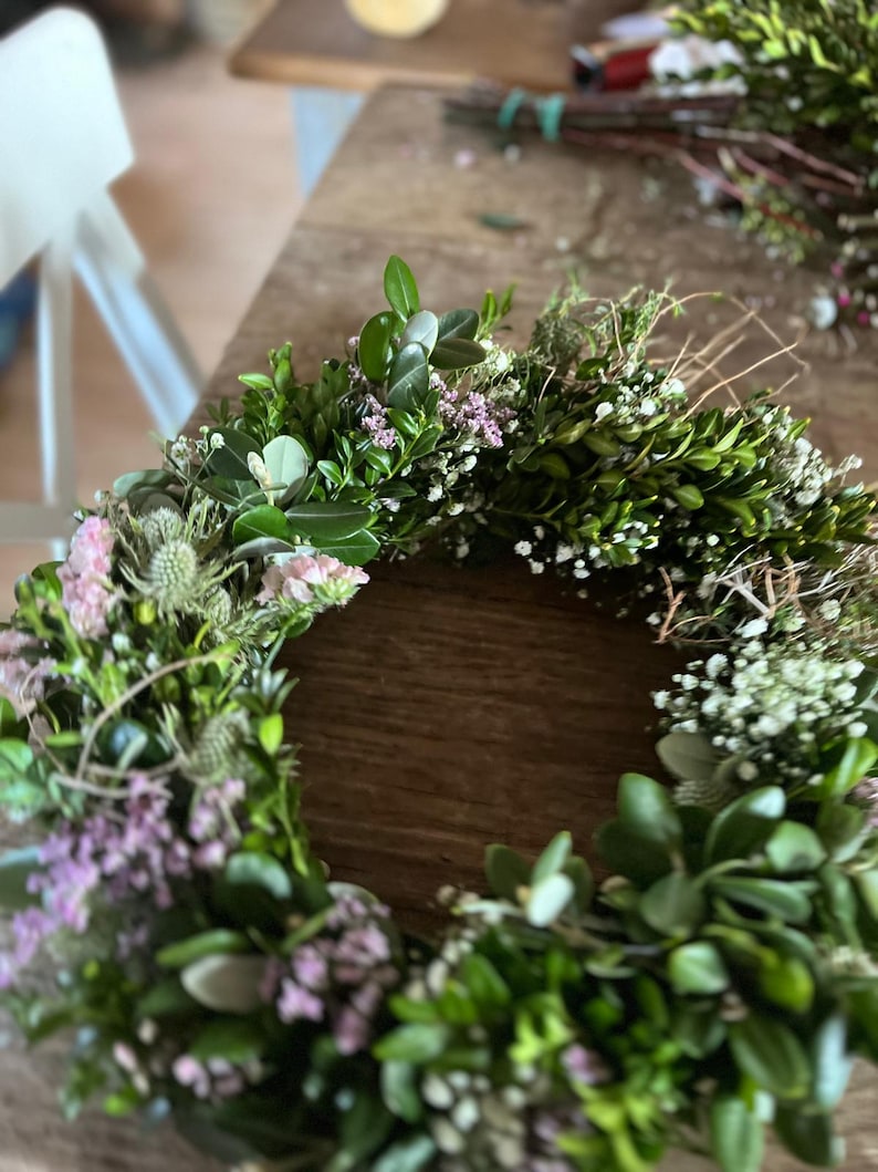 May include: A circular wreath constructed from green boxwood leaves, interspersed with small clusters of white baby's breath, light pink statice flowers, and spiky blue-green thistle heads, arranged in a dense, textured botanical ring.