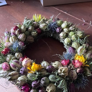 Könnte beinhalten: Ein kreisförmiger Kranz aus getrockneten botanischen Elementen, bestehend aus einer dichten Anordnung von grünen Samenkapseln, stacheligen blauen Distelköpfen und kleinen, leuchtenden Blüten in Gelb-, Magenta- und Dunkellila-Tönen.