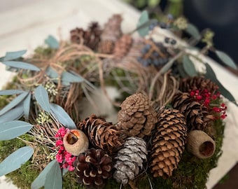 May include: A circular wreath constructed from a woven twig base covered in green moss. The wreath is decorated with various brown pinecones, light blue eucalyptus leaves, small clusters of bright pink flowers, and acorn caps.