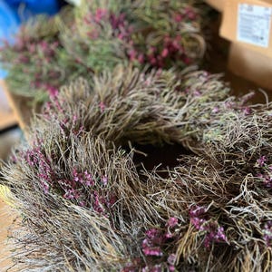 K&ouml;nnte beinhalten: Ein kreisf&ouml;rmiger Kranz aus getrockneten, d&uuml;nnen, braunen Zweigen und &Auml;sten, der mit kleinen B&uuml;scheln leuchtend violetter Bl&uuml;ten verziert ist, die &uuml;ber die gesamte Struktur verteilt sind. Der Kranz hat ein strukturiertes, nat&uuml;rliches Aussehen.