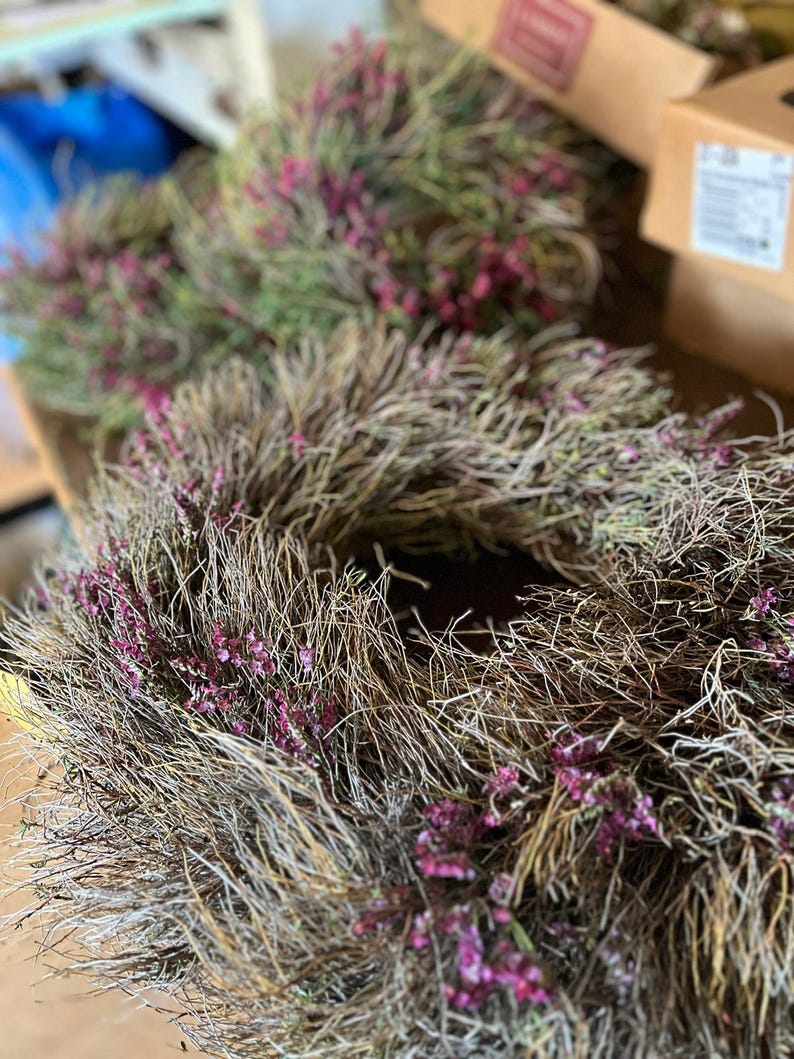 K&ouml;nnte beinhalten: Ein kreisf&ouml;rmiger Kranz aus getrockneten, d&uuml;nnen, braunen Zweigen und &Auml;sten, der mit kleinen B&uuml;scheln leuchtend violetter Bl&uuml;ten verziert ist, die &uuml;ber die gesamte Struktur verteilt sind. Der Kranz hat ein strukturiertes, nat&uuml;rliches Aussehen.