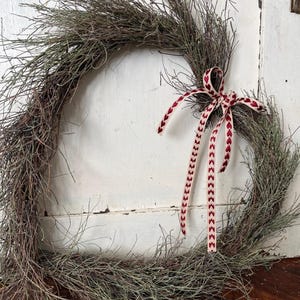 Puede incluir: Una corona r&uacute;stica de ramas con una cinta roja y blanca atada en un lazo. La corona est&aacute; hecha de ramas naturales y es perfecta para un estilo de decoraci&oacute;n de granja o de campo.