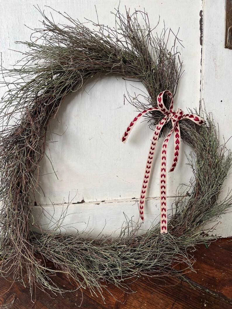 Puede incluir: Una corona r&uacute;stica de ramas con una cinta roja y blanca atada en un lazo. La corona est&aacute; hecha de ramas naturales y es perfecta para un estilo de decoraci&oacute;n de granja o de campo.