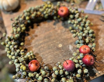 Puede incluir: Una corona circular construida con numerosas bayas verdes pequeñas y texturizadas, intercaladas con varias manzanas rojas, redondas y lisas. La corona está dispuesta sobre una superficie de madera oscura y desgastada, mostrando una composición botánica natural.