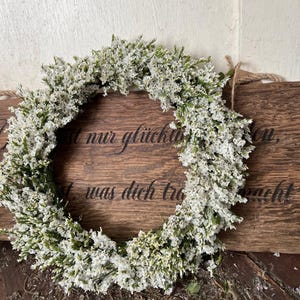 May include: A circular wreath of white flowers and green leaves. The wreath is displayed on a wooden sign with German text. The sign is attached to a light-coloured wooden wall.