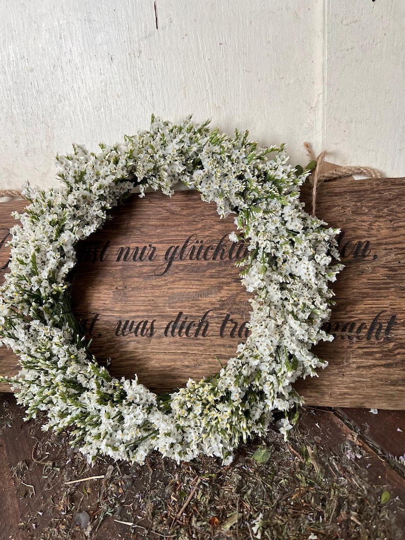 May include: A circular wreath of white flowers and green leaves. The wreath is displayed on a wooden sign with German text. The sign is attached to a light-coloured wooden wall.