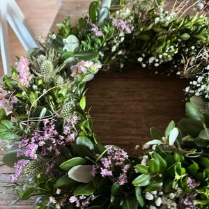 May include: A handmade wreath featuring a variety of greenery, including boxwood, accented with small white and purple flowers. The wreath is arranged in a circular shape, perfect for seasonal decor.