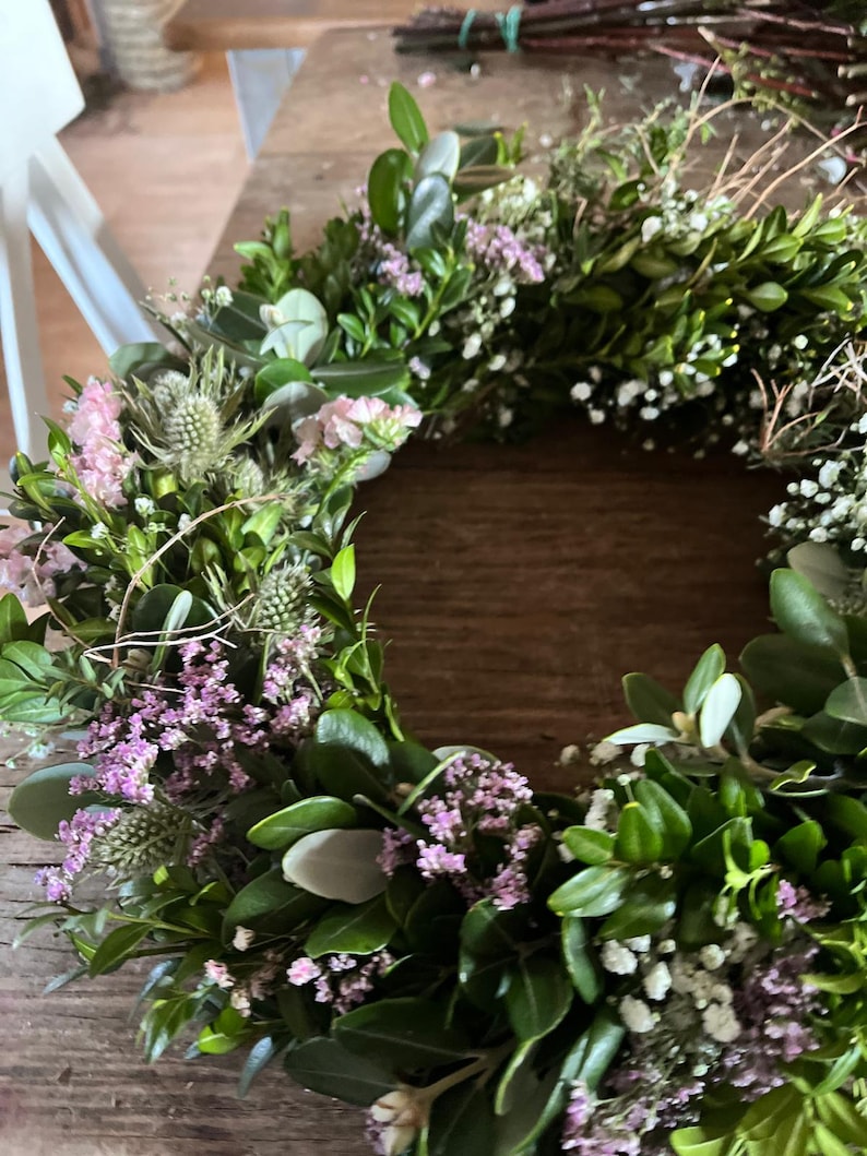 May include: A handmade wreath featuring a variety of greenery, including boxwood, accented with small white and purple flowers. The wreath is arranged in a circular shape, perfect for seasonal decor.