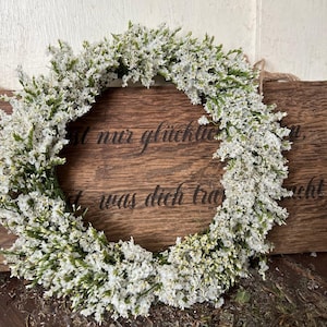 May include: A circular wreath of small white flowers and green stems. The wreath is set against a wooden background with etched text. A natural, decorative wreath.