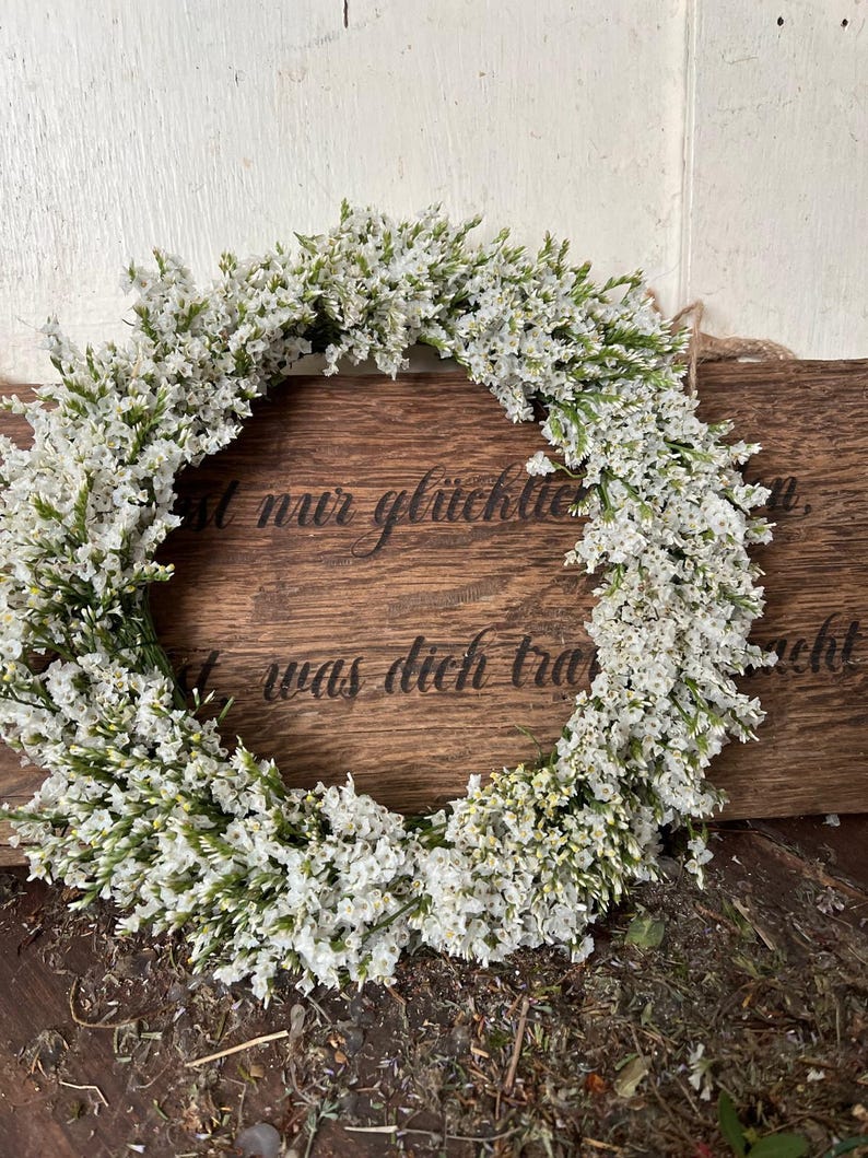 May include: A circular wreath of small white flowers and green stems. The wreath is set against a wooden background with etched text. A natural, decorative wreath.