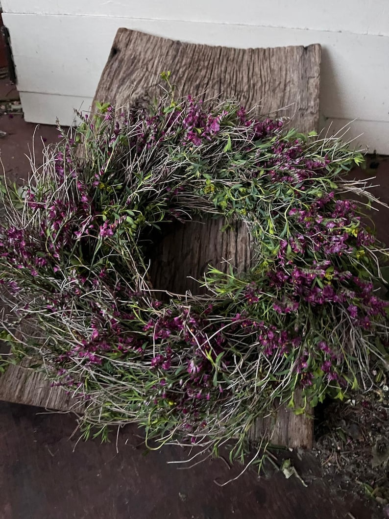 K&ouml;nnte beinhalten: Ein kreisf&ouml;rmiger Kranz aus d&uuml;nnen, getrockneten braunen Zweigen und St&auml;ngeln, dicht verwoben mit kleinen, leuchtend violetten Bl&uuml;ten und d&uuml;nnen gr&uuml;nen Bl&auml;ttern, der ein strukturiertes, nat&uuml;rliches und abgerundetes botanisches Arrangement bildet.