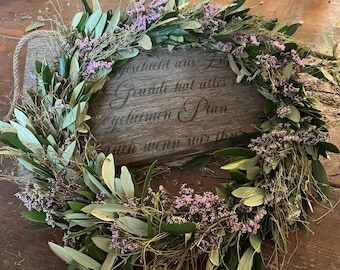 Puede incluir: Una corona hecha a mano con ramas de olivo, pequeñas flores moradas y hierbas secas. La corona está dispuesta alrededor de un letrero de madera con texto grabado. La corona se exhibe sobre una mesa de madera.