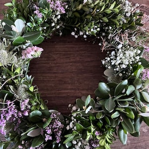 May include: A circular wreath constructed from dark green boxwood leaves, interspersed with small clusters of white baby's breath, light purple statice flowers, and spiky green thistle heads, creating a dense and textured botanical arrangement.