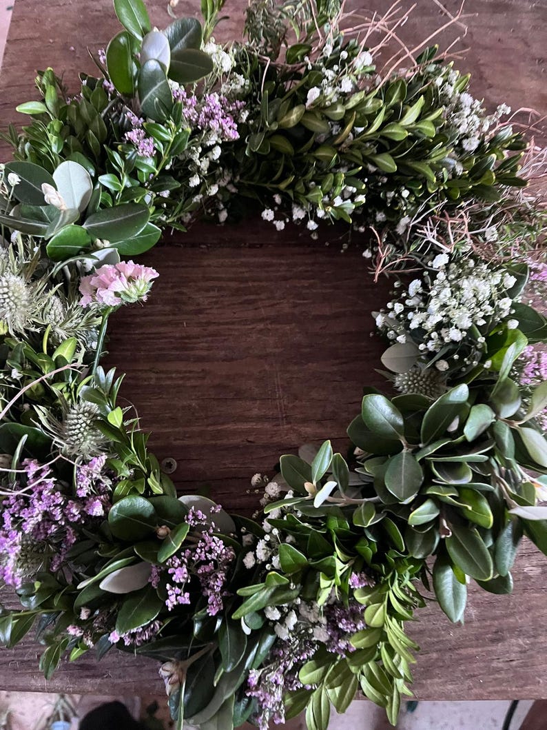 May include: A circular wreath constructed from dark green boxwood leaves, interspersed with small clusters of white baby's breath, light purple statice flowers, and spiky green thistle heads, creating a dense and textured botanical arrangement.