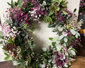 May include: A circular wreath composed of various green leaves, accented with clusters of small, vibrant pink and white flowers. The wreath is displayed on a wooden surface, creating a natural and rustic aesthetic. The wreath is a decorative item.