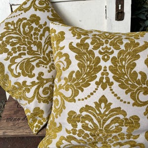 May include: Two square cushions featuring a white backdrop and a gold damask design. The cushions are made from a textured material and are positioned on a wooden surface. These cushions are a decorative home decor item.