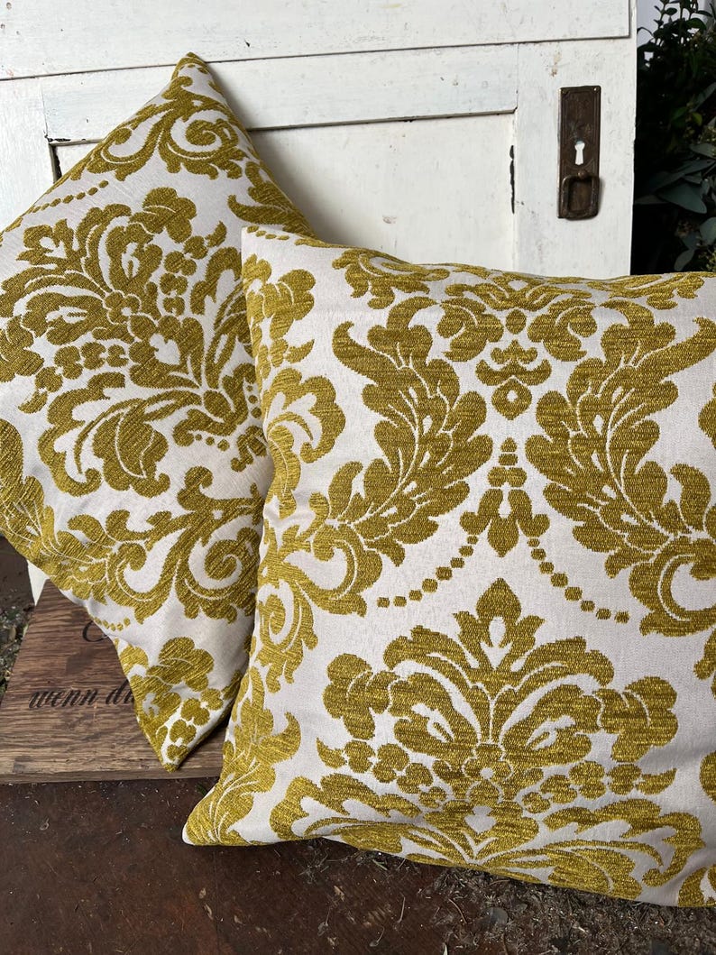 May include: Two square cushions featuring a white backdrop and a gold damask design. The cushions are made from a textured material and are positioned on a wooden surface. These cushions are a decorative home decor item.