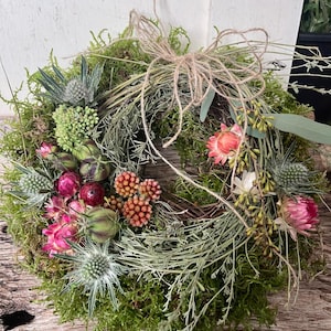 Könnte beinhalten: Ein runder Kranz aus grünem Moos, verziert mit getrockneten Disteln, rosa Strohblumen und kleinen Beerenclustern. Das Arrangement ist mit einer dünnen, hellbraunen Schnur akzentuiert, die oben zu einer Schleife gebunden ist und verschiedene getrocknete Gräser enthält.