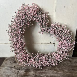 Könnte beinhalten: Ein herzförmiger Kranz aus kleinen, hellrosa Blüten. Der Kranz ist vor einer rustikalen, verwitterten Holzoberfläche und einem weißen Hintergrund platziert. Die Blüten sind dicht gepackt und erzeugen ein volles, strukturiertes Aussehen.