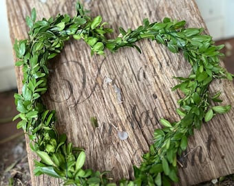 May include: A heart-shaped wreath crafted from fresh green boxwood leaves. The wreath sits on a weathered wooden surface. The leaves are a vibrant green, creating a natural design. The wooden surface has faint text.