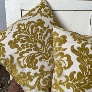 May include: Two square cushions featuring a white backdrop and a gold damask design. The cushions are made from a textured material and are placed on a wooden surface. The cushions have a classic, elegant design.