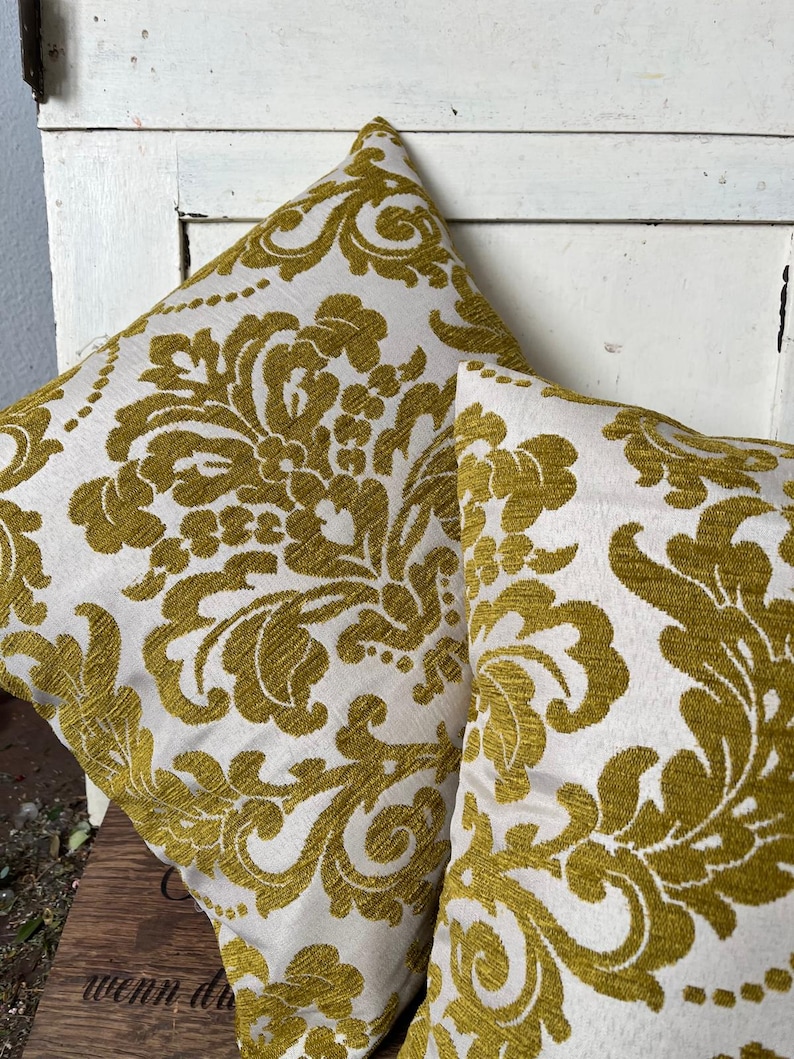 May include: Two square cushions featuring a white backdrop and a gold damask design. The cushions are made from a textured material and are placed on a wooden surface. The cushions have a classic, elegant design.