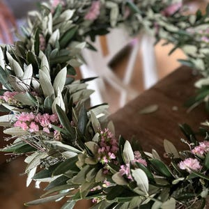 Puede incluir: Primer plano de una corona hecha a mano con ramas de olivo y peque&ntilde;as flores rosas. La corona circular descansa sobre una superficie de madera. Las hojas son verdes y gris plateado.