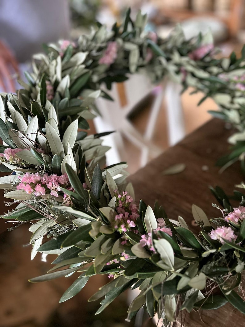 Puede incluir: Primer plano de una corona hecha a mano con ramas de olivo y peque&ntilde;as flores rosas. La corona circular descansa sobre una superficie de madera. Las hojas son verdes y gris plateado.