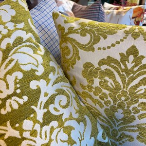 May include: Close-up of decorative cushions with an olive green and white floral damask pattern. The cushions are made of a textured fabric and are arranged in a display, showcasing home decor.