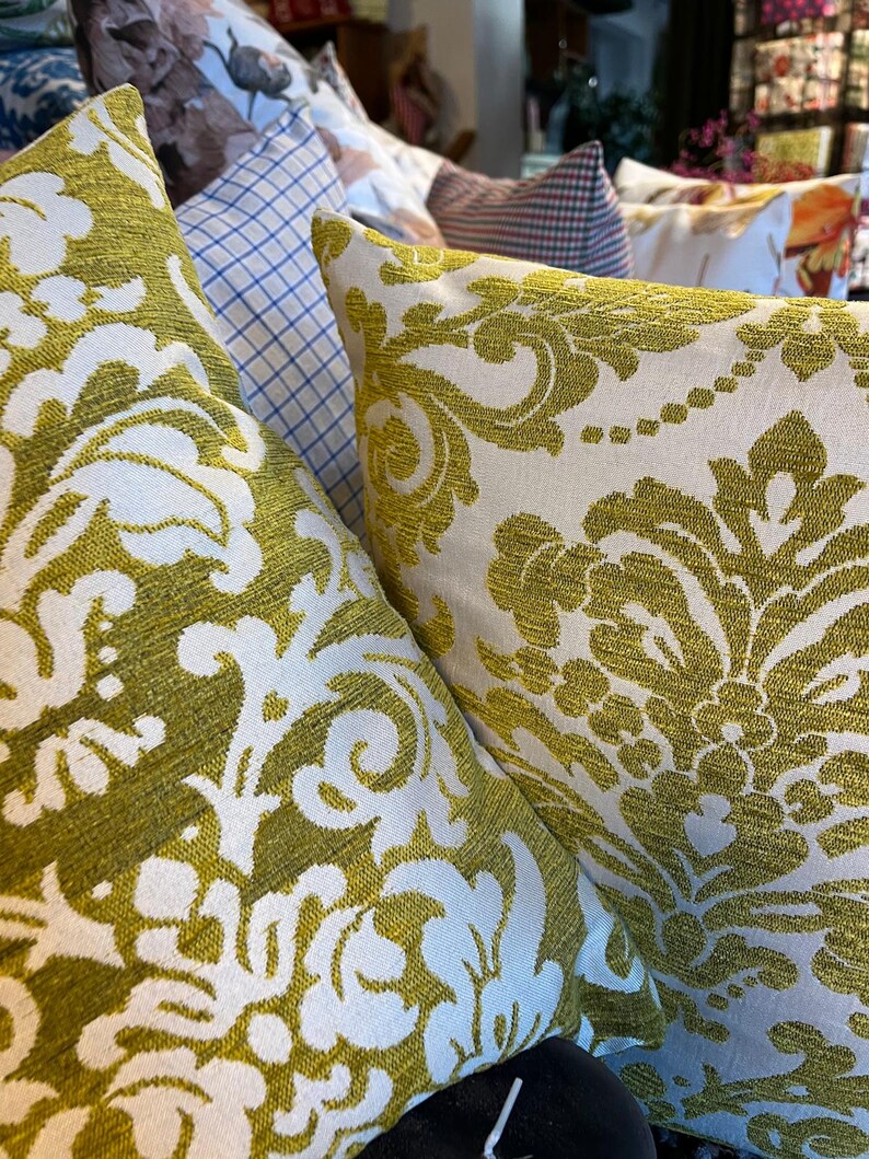 May include: Close-up of decorative cushions with an olive green and white floral damask pattern. The cushions are made of a textured fabric and are arranged in a display, showcasing home decor.