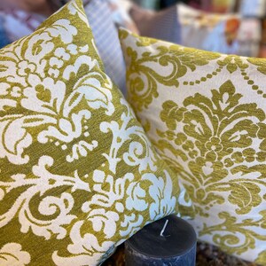 May include: Two cushions with an olive green and white damask pattern. A dark grey candle is in front of the cushions. Other cushions are visible in the background, creating a cozy atmosphere.