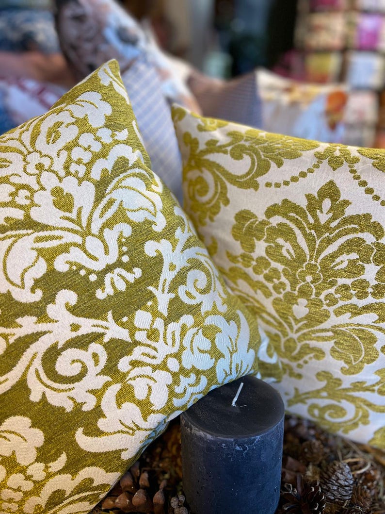May include: Two cushions with an olive green and white damask pattern. A dark grey candle is in front of the cushions. Other cushions are visible in the background, creating a cozy atmosphere.
