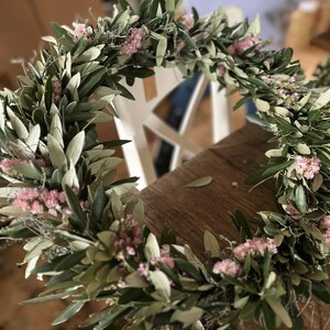 Puede incluir: Una corona hecha a mano con ramas de olivo y peque&ntilde;as flores rosas. La corona est&aacute; dispuesta en forma circular, con las hojas verdes y las flores rosas creando un aspecto natural y org&aacute;nico. Perfecta para la decoraci&oacute;n del hogar o eventos especiales.