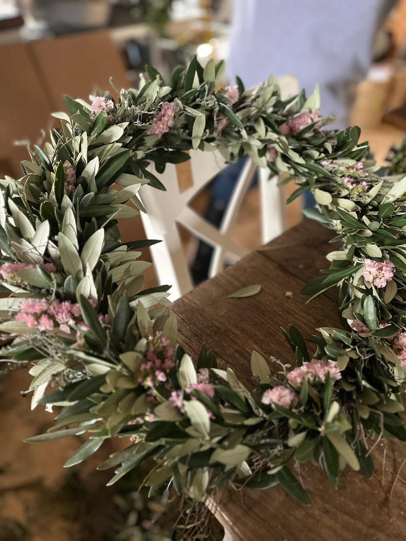Puede incluir: Una corona hecha a mano con ramas de olivo y peque&ntilde;as flores rosas. La corona est&aacute; dispuesta en forma circular, con las hojas verdes y las flores rosas creando un aspecto natural y org&aacute;nico. Perfecta para la decoraci&oacute;n del hogar o eventos especiales.