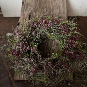 K&ouml;nnte beinhalten: Ein kreisf&ouml;rmiger Kranz aus getrockneten, d&uuml;nnen, holzigen Zweigen und St&auml;ngeln, dicht durchsetzt mit kleinen, leuchtend violett-rosa Bl&uuml;ten und feinem gr&uuml;nen Blattwerk, was ein strukturiertes, nat&uuml;rliches und organisches Erscheinungsbild erzeugt.