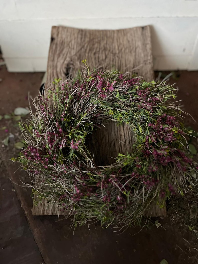 K&ouml;nnte beinhalten: Ein kreisf&ouml;rmiger Kranz aus getrockneten, d&uuml;nnen, holzigen Zweigen und St&auml;ngeln, dicht durchsetzt mit kleinen, leuchtend violett-rosa Bl&uuml;ten und feinem gr&uuml;nen Blattwerk, was ein strukturiertes, nat&uuml;rliches und organisches Erscheinungsbild erzeugt.