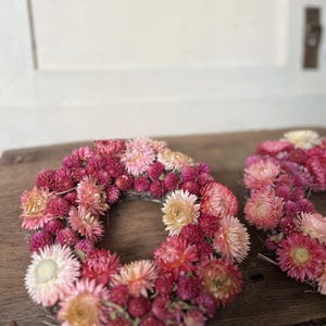 May include: A handmade floral wreath composed of dried flowers in shades of pink, red, and cream. The wreath is circular and rests on a wooden surface. The flowers are densely packed, creating a full and textured appearance.
