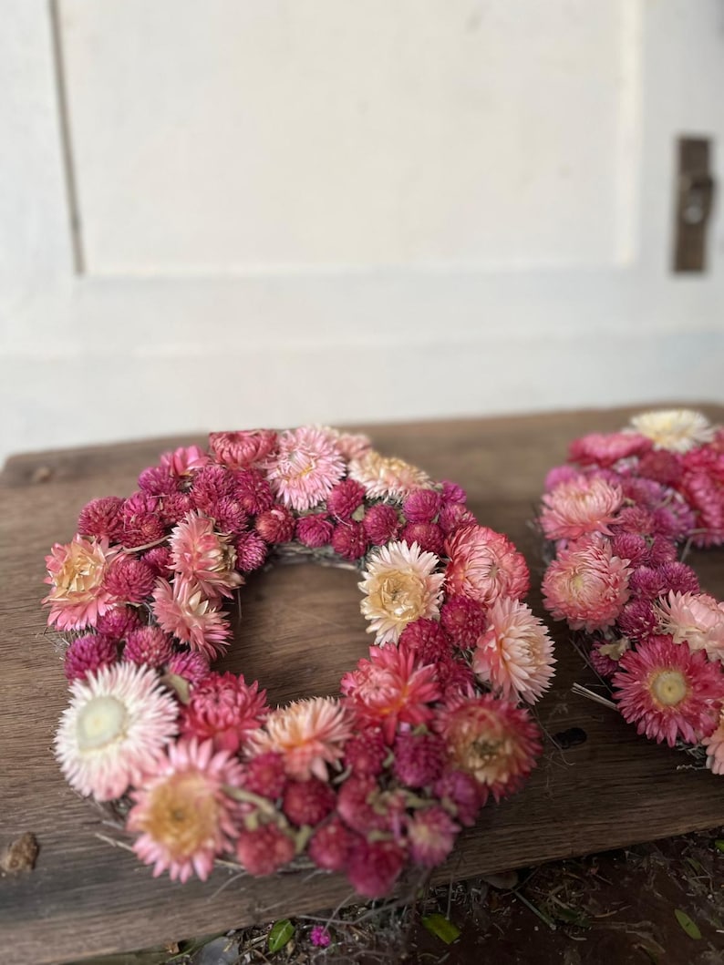 May include: A handmade floral wreath composed of dried flowers in shades of pink, red, and cream. The wreath is circular and rests on a wooden surface. The flowers are densely packed, creating a full and textured appearance.