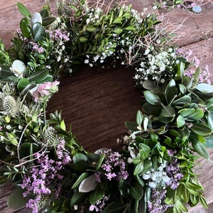 May include: A circular wreath constructed from dense green boxwood leaves, interspersed with small clusters of white baby's breath, light purple statice flowers, and spiky blue-green thistle heads, creating a textured botanical arrangement.