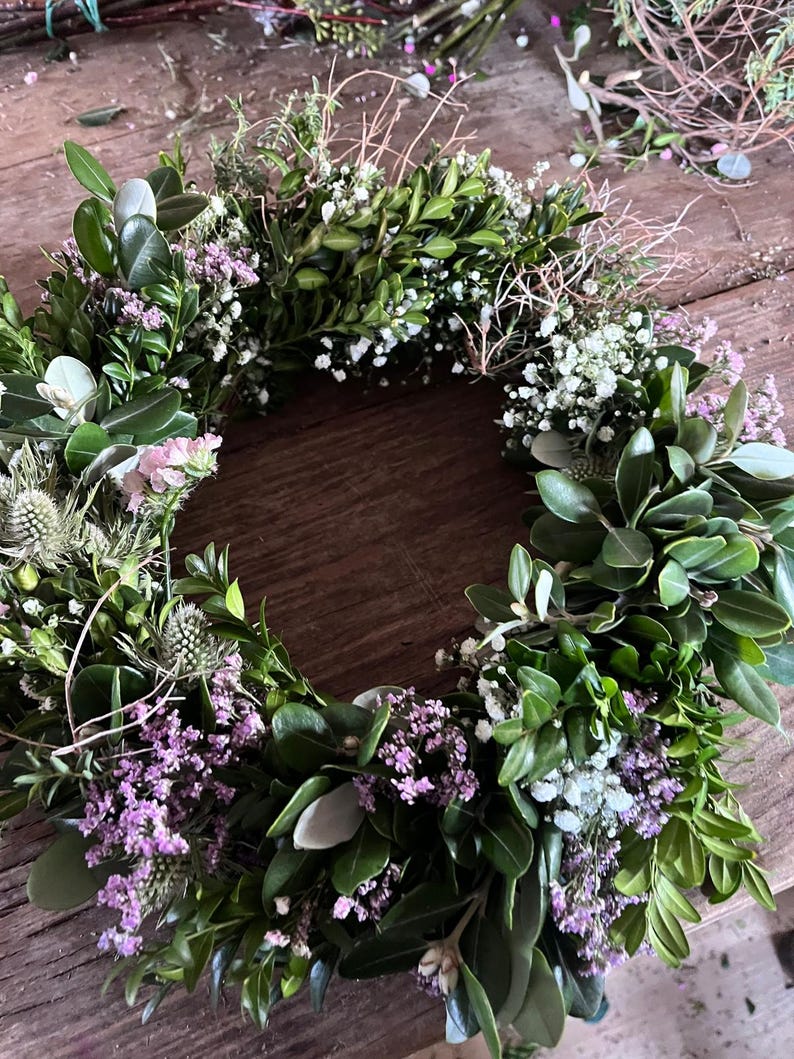 May include: A circular wreath constructed from dense green boxwood leaves, interspersed with small clusters of white baby's breath, light purple statice flowers, and spiky blue-green thistle heads, creating a textured botanical arrangement.