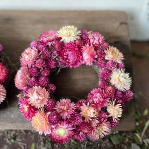 May include: A circular floral wreath composed of various shades of pink and cream-colored dried flowers. The wreath is arranged in a dense, full circle, with a mix of textures and sizes. The flowers are set against a natural, neutral background.